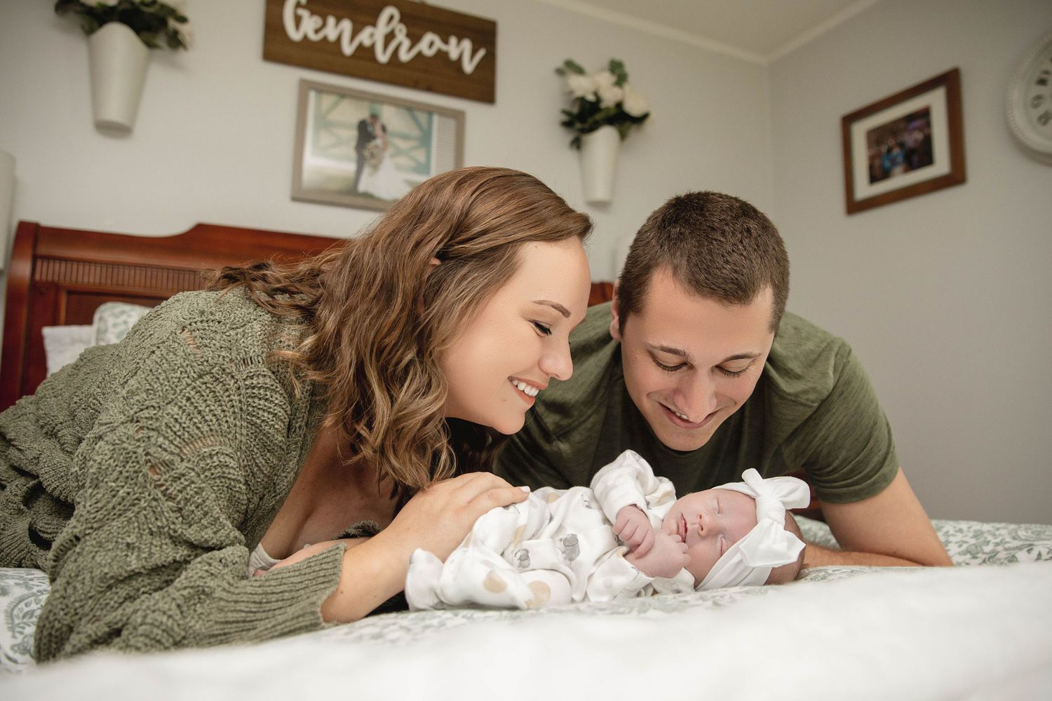 Parents lie on bed looking adoringly at their newborn baby under a wall sign.