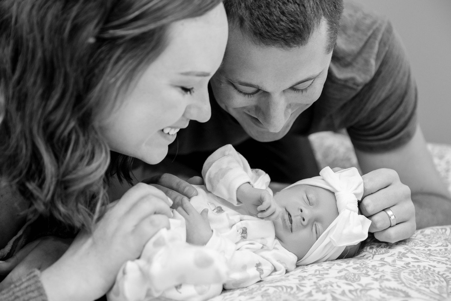 A black and white image capturing parents admiring their newborn baby on bed linens.