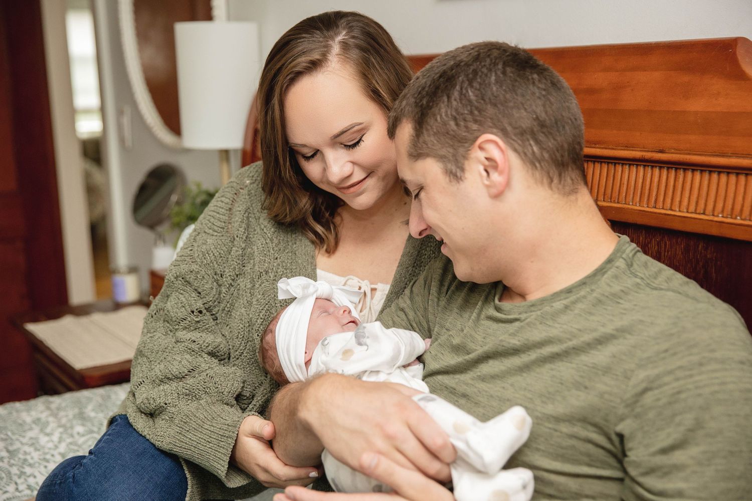 Parents snuggle with their newborn baby while sitting on a bed near a wooden headboard.