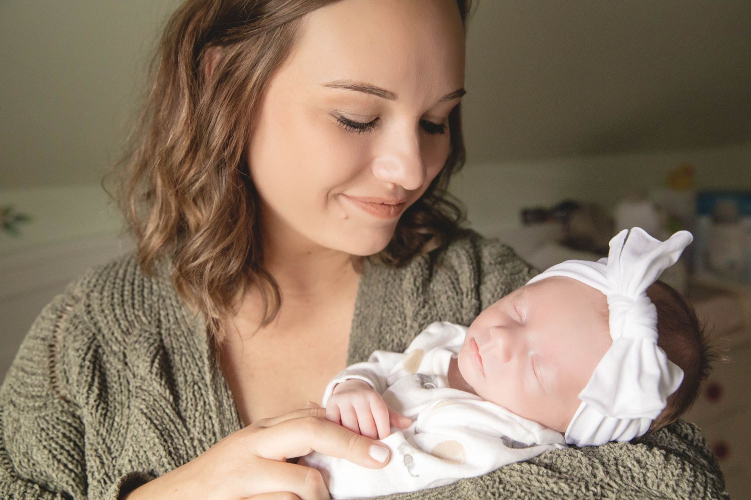 A mother in a gray sweater gazes lovingly at her newborn baby wrapped in white.