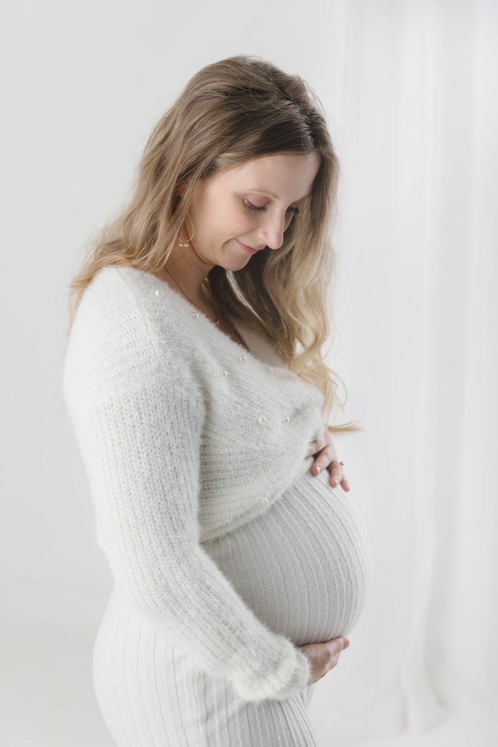 An intimate maternity portrait featuring an expectant mother in a white sweater against a light background.