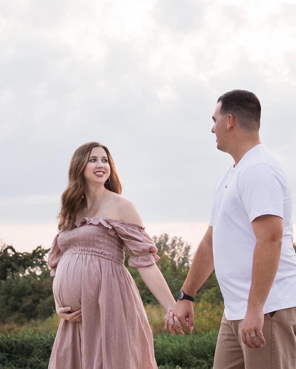 A romantic outdoor maternity photo featuring a couple in casual attire holding hands at sunset.