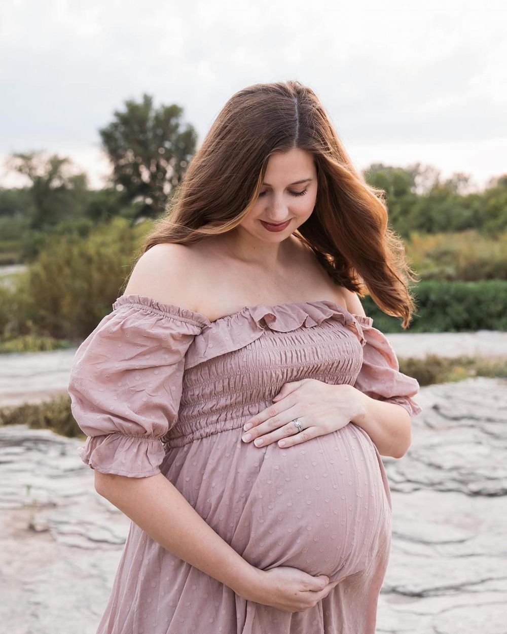 Maternity photo of woman in dusty rose off-shoulder dress touching her baby bump.