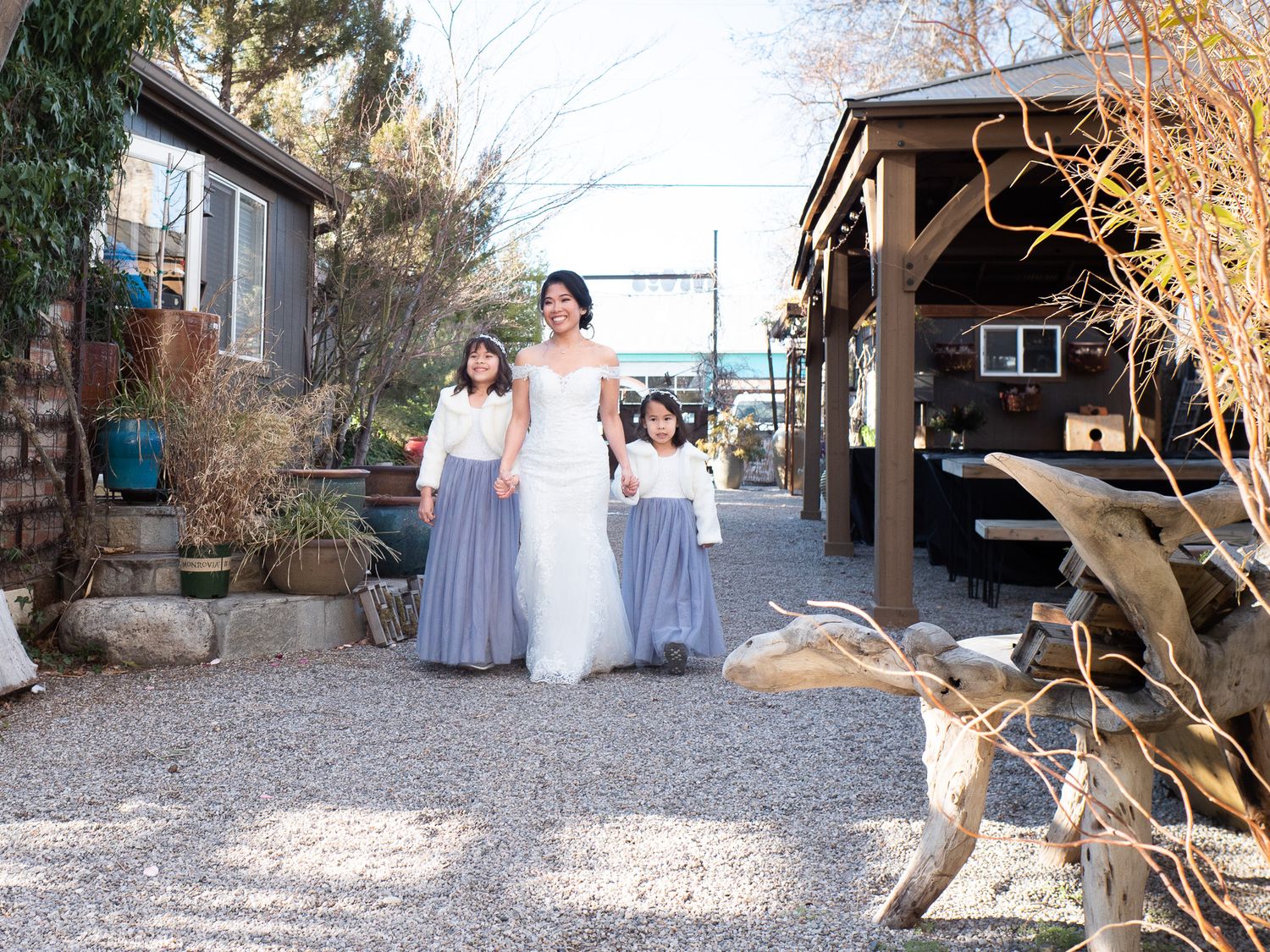 Bride in formal attire with her daughters stand together on a gravel path beside a covered patio area at Sierra Water Gardens.