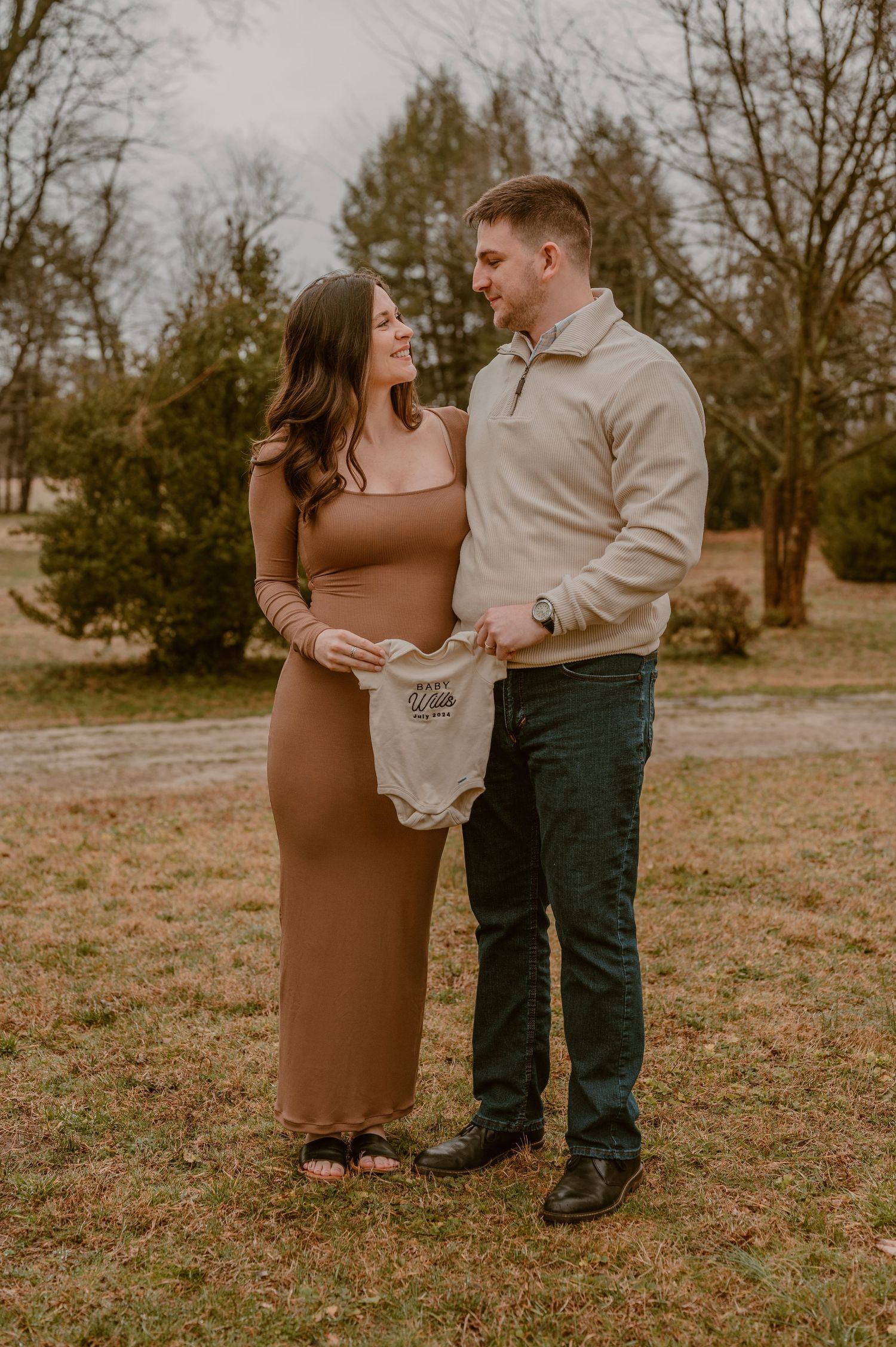 A couple stands together in a field holding a baby onesie in a romantic maternity photo. photo by Samantha Campbell imagery