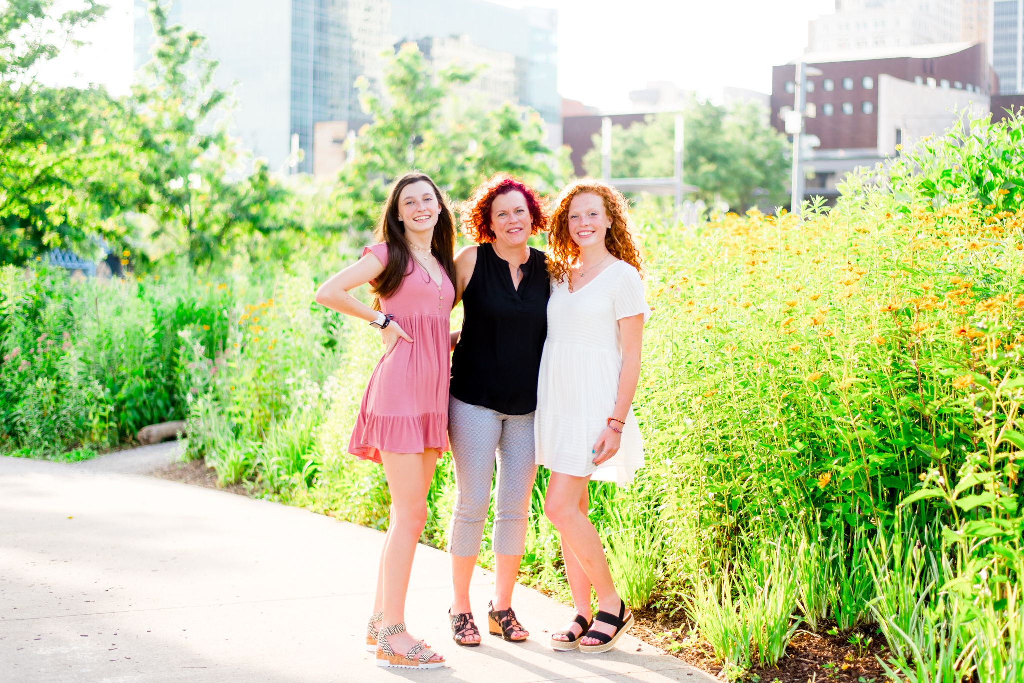 mom and two teenage daughters smiling in Smale Park in front of flowers and buildings