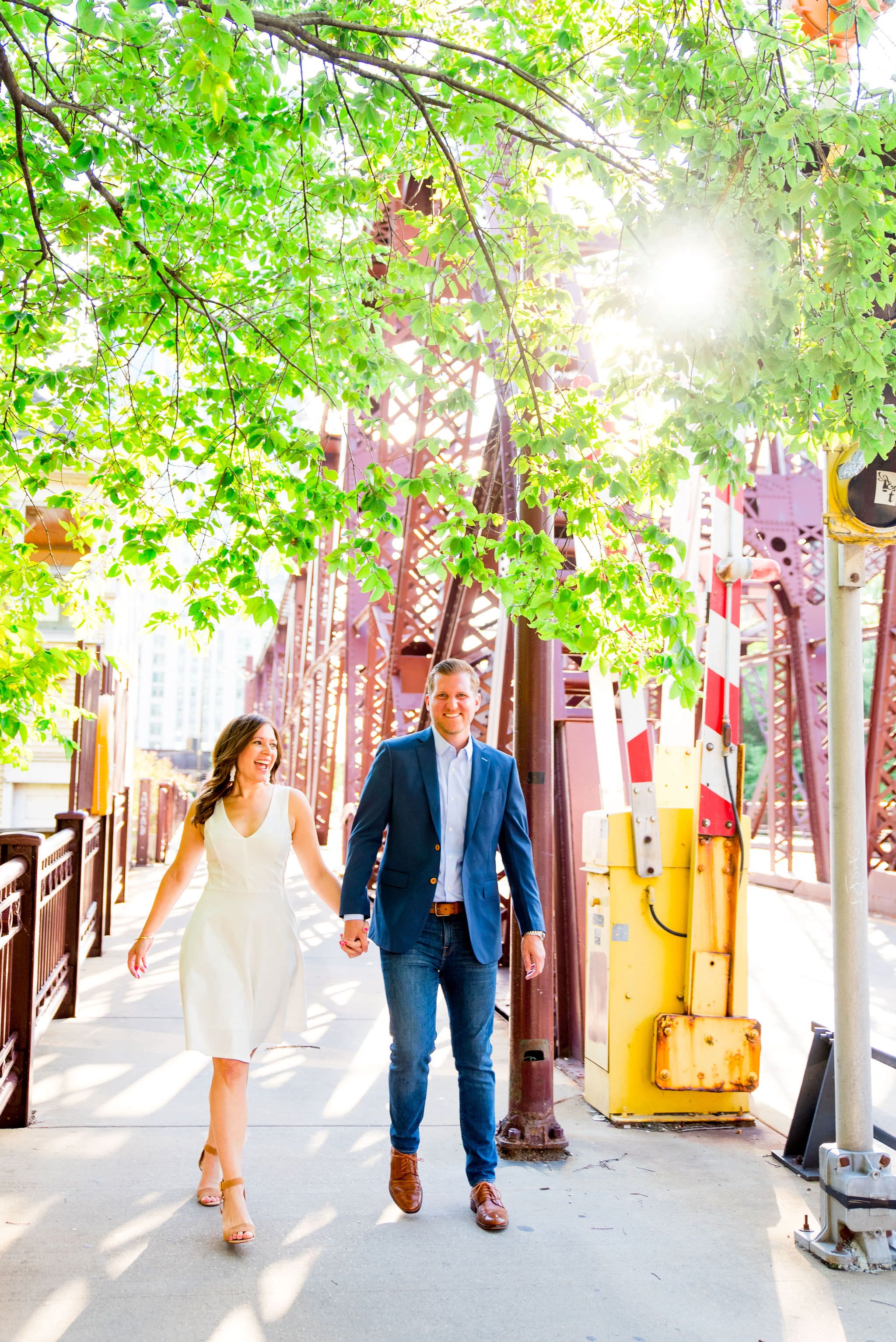 woman in white dress and man in navy suit walking by Kinzie St Bridge in Chicago for sunset engagement pictures