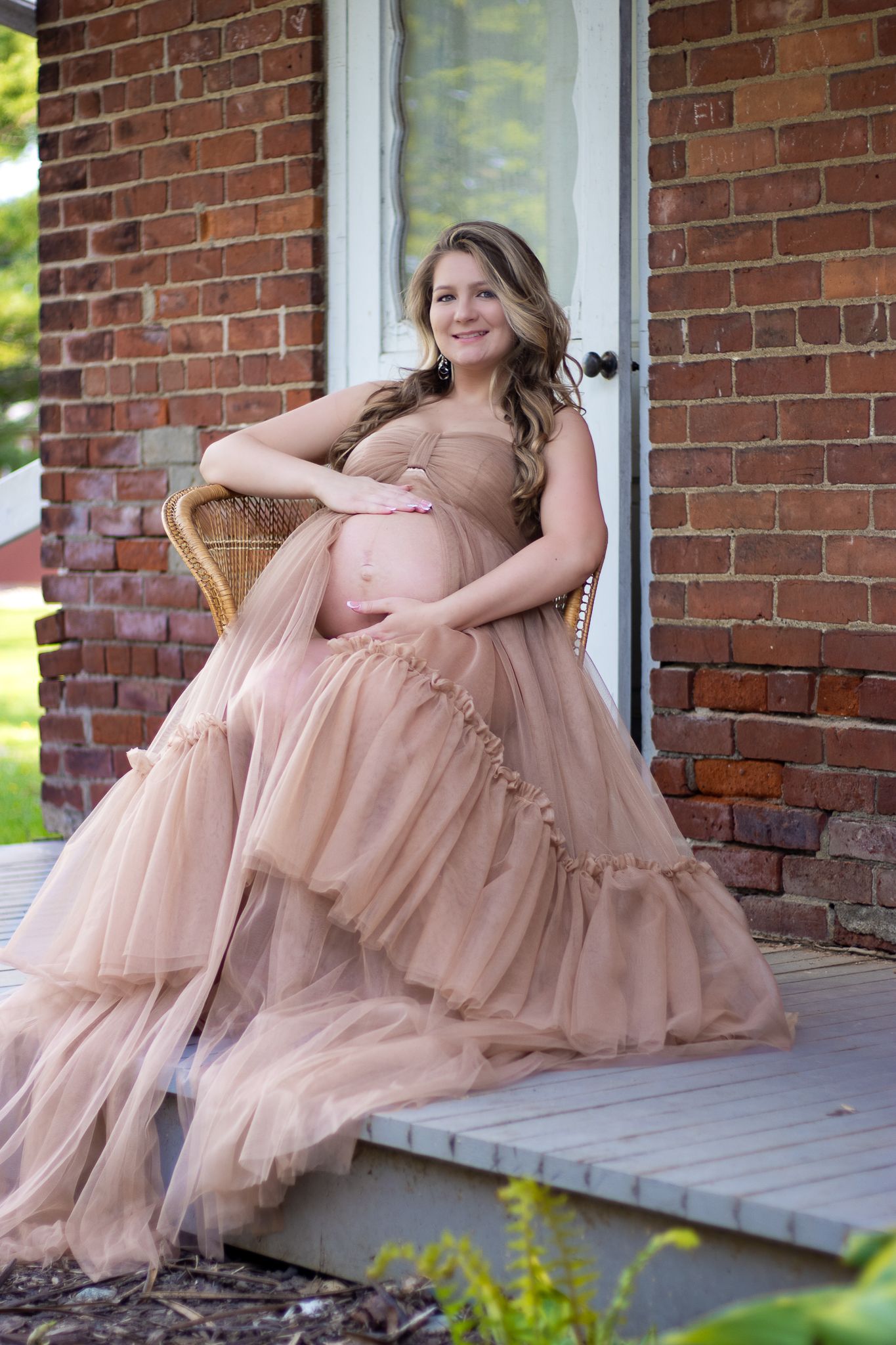 A soft blush tulle maternity gown photographed on a rustic porch with brick walls.