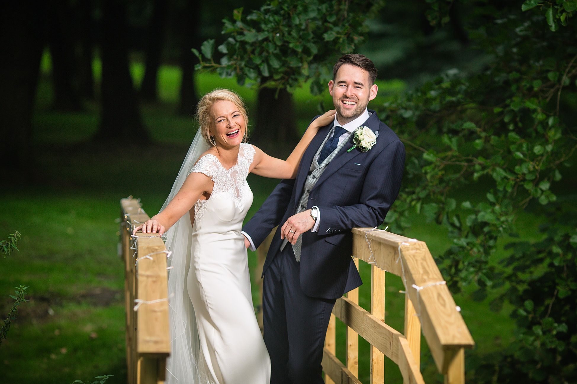 Bride and groom stand laughing on a wooden bridge in the garden of their Cheshire Marquee wedding