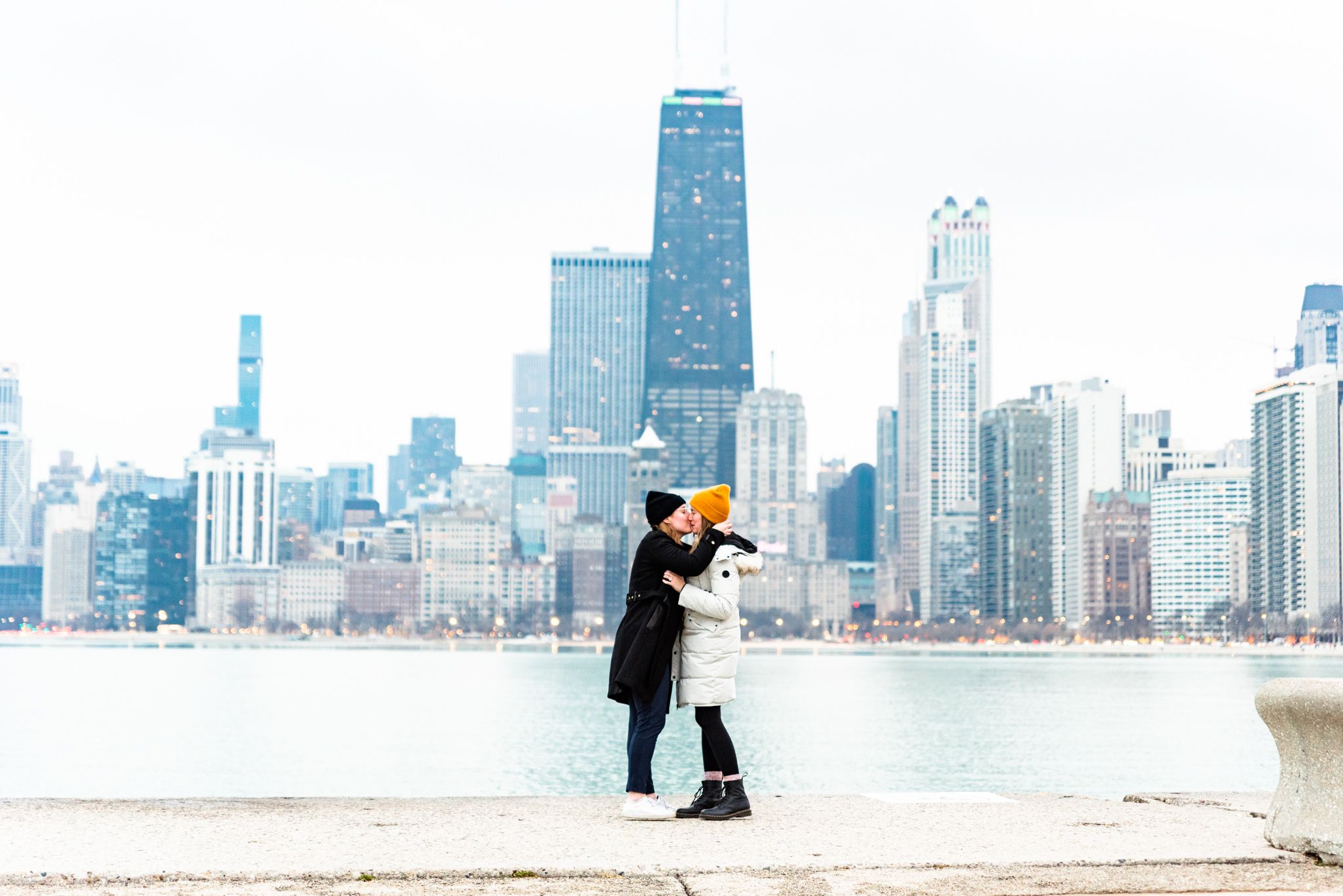 woman in black coat and woman in white coat kissing on pier at North Avenue Beach Chicago after proposal in Chicago
