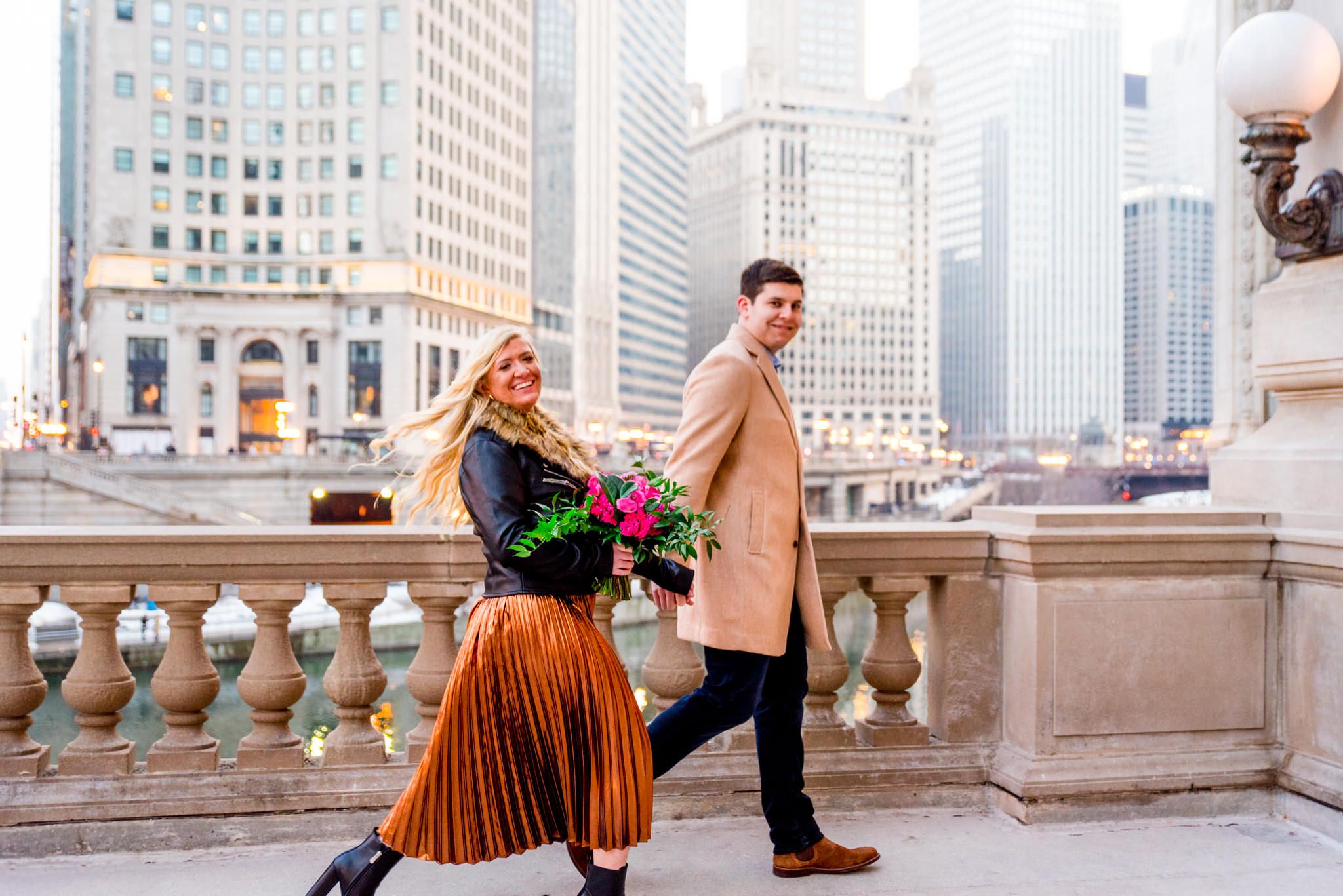man in tan coat walking with woman in copper skirt in front of Wrigley Building engagement photos