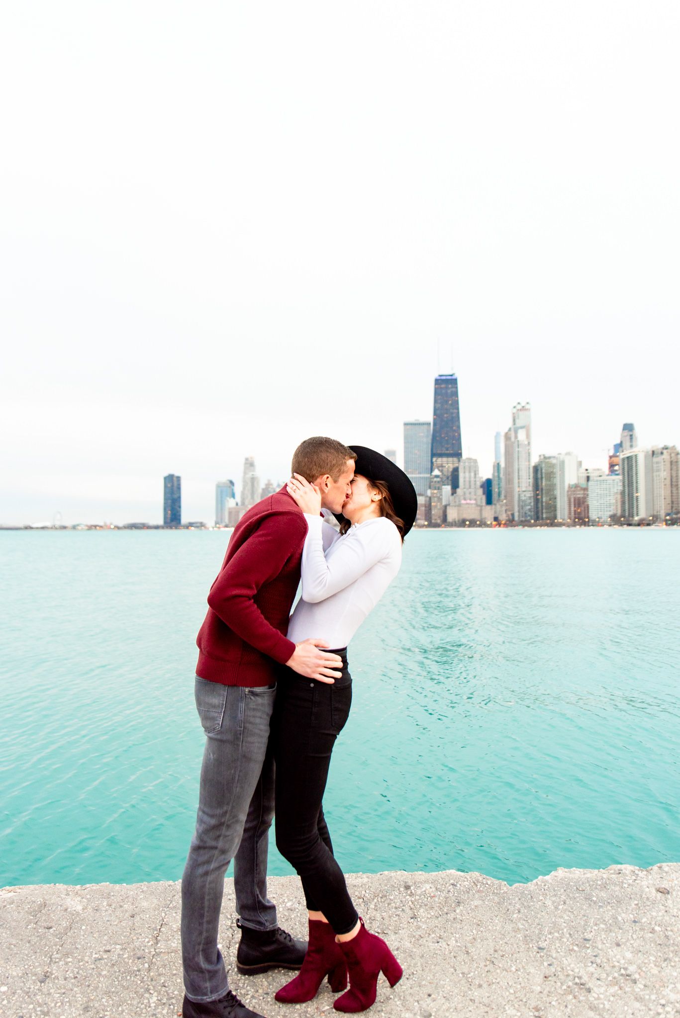 man in red sweater kissing woman in white shirt and black hat kissing at North Avenue Beach pier engagement pictures