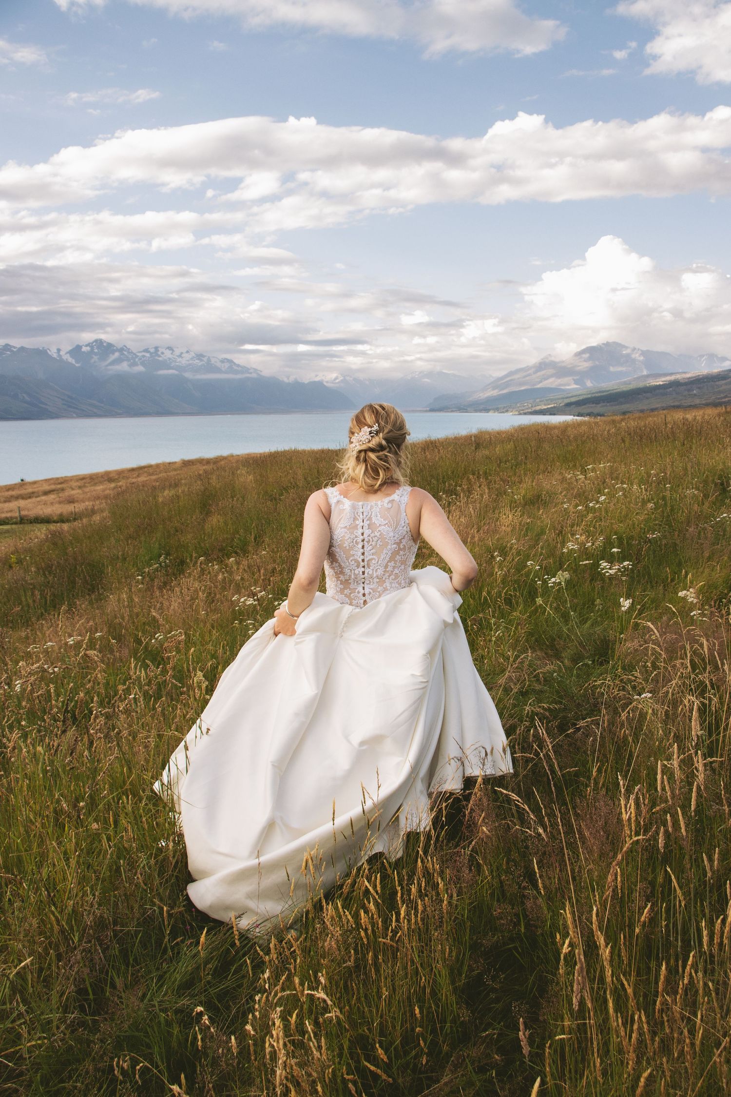 Bride in flowing white dress runs through mountain meadow with lake and mountains in background.