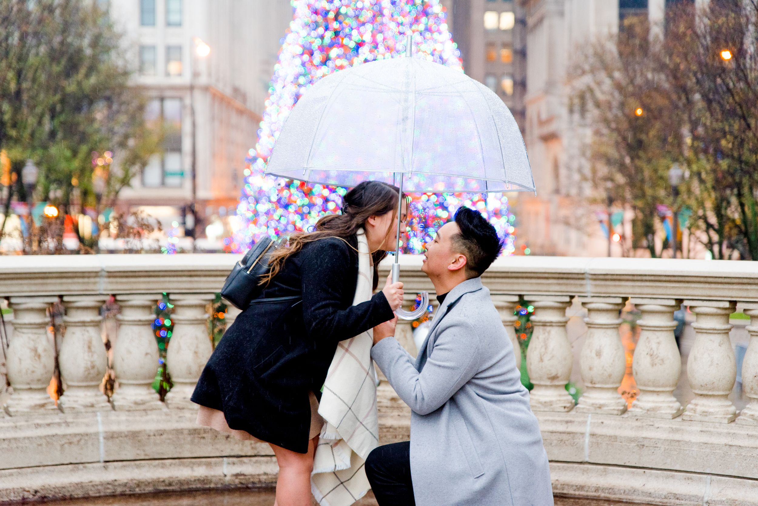 man in grey coat kneeling proposing to woman in black coat and clear umbrella in front of Millennium Park Christmas Tree