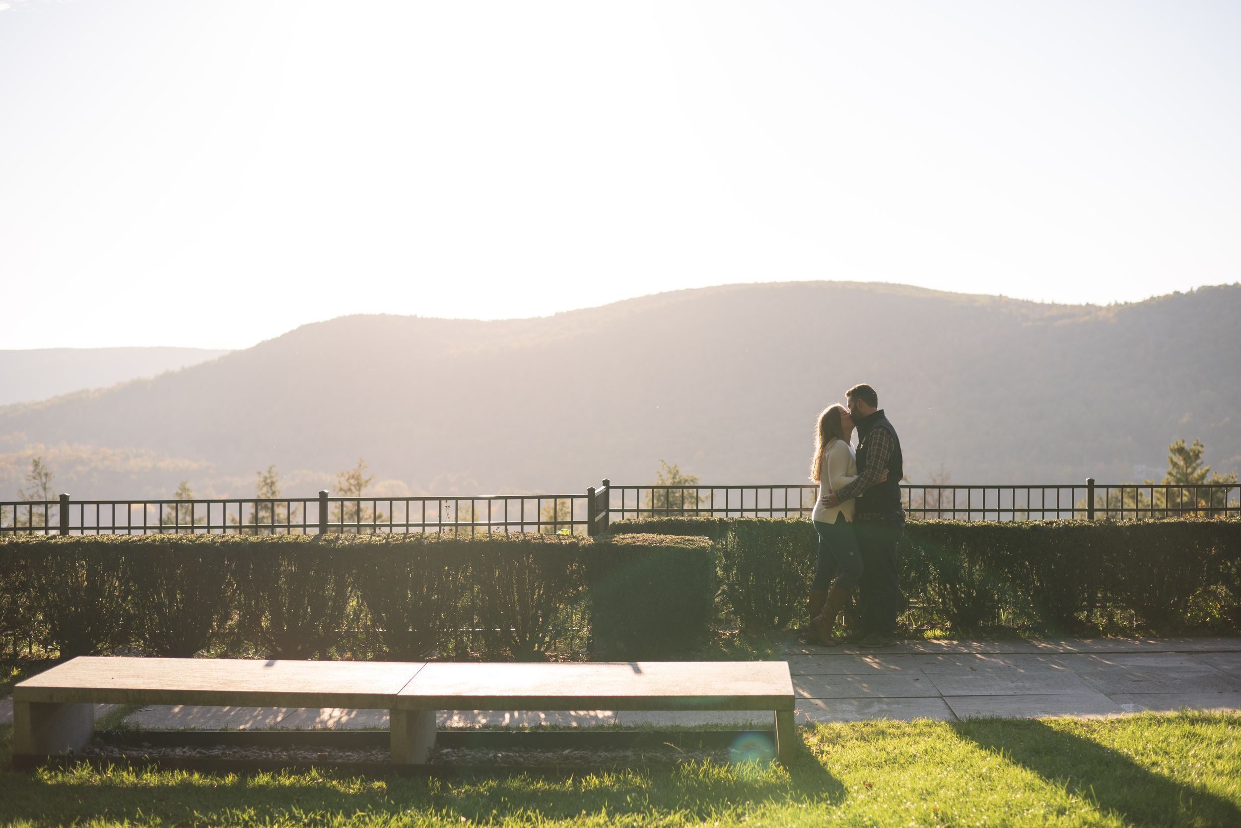A silhouetted couple stands on a scenic overlook at sunset with mountains in the distance.