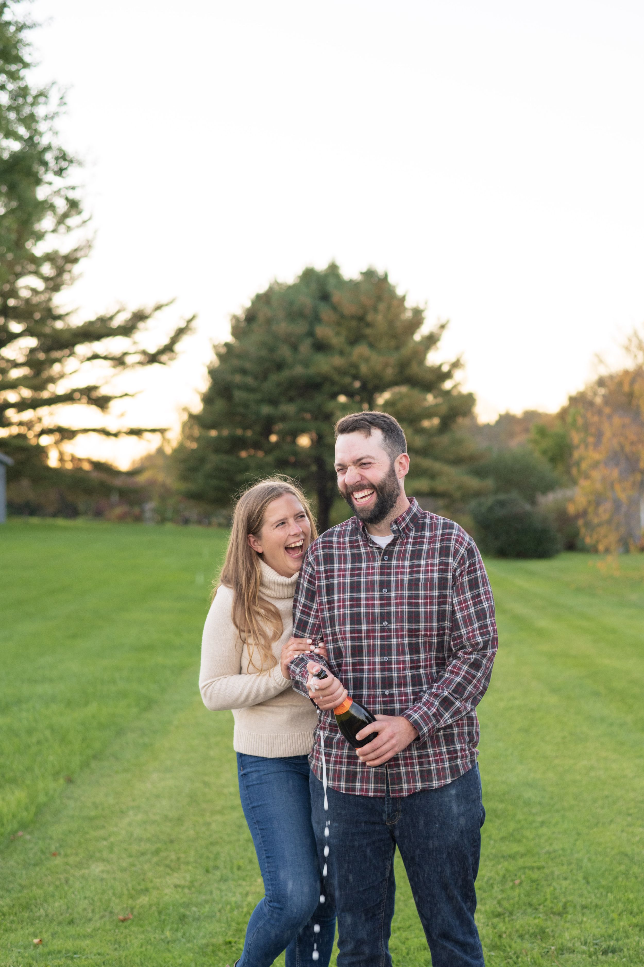 Joyful couple laughing while popping champagne in a sunlit garden.