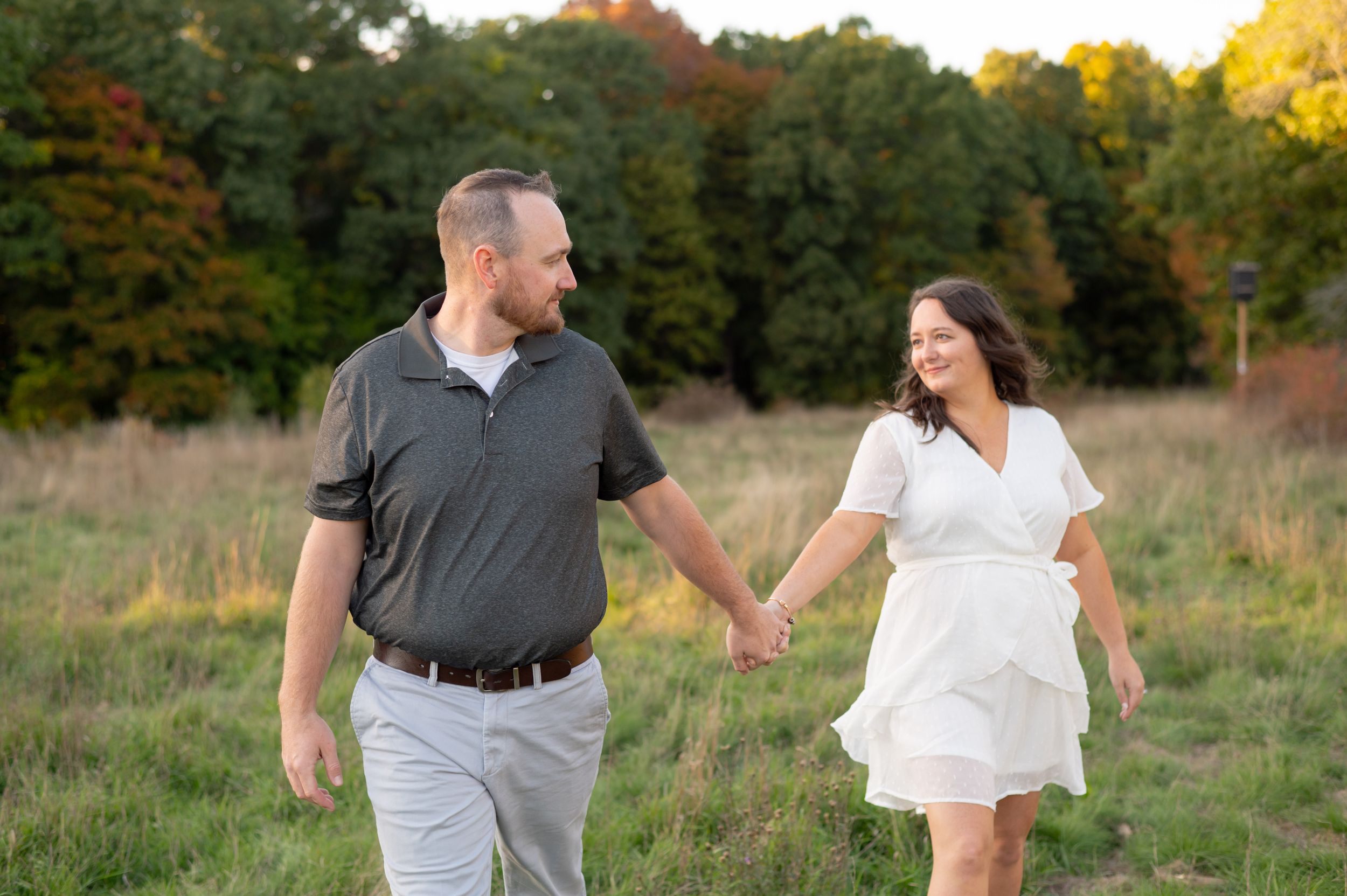 A couple walks hand in hand through a fall meadow with golden trees in the background.