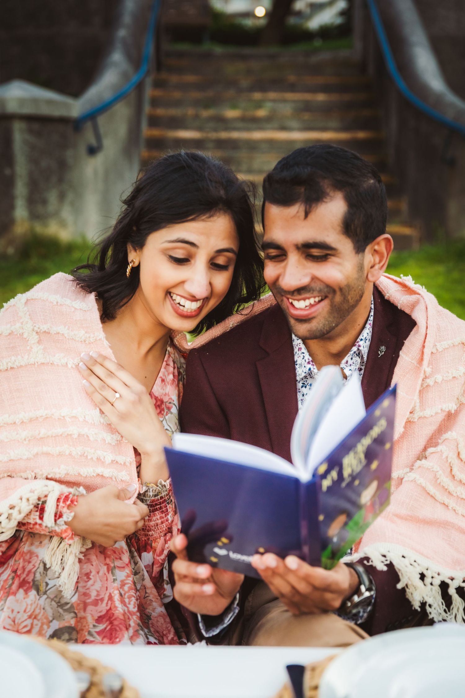 Indian couple smiling reading a book together outside wraped in a blanket for a pre wedding shoot