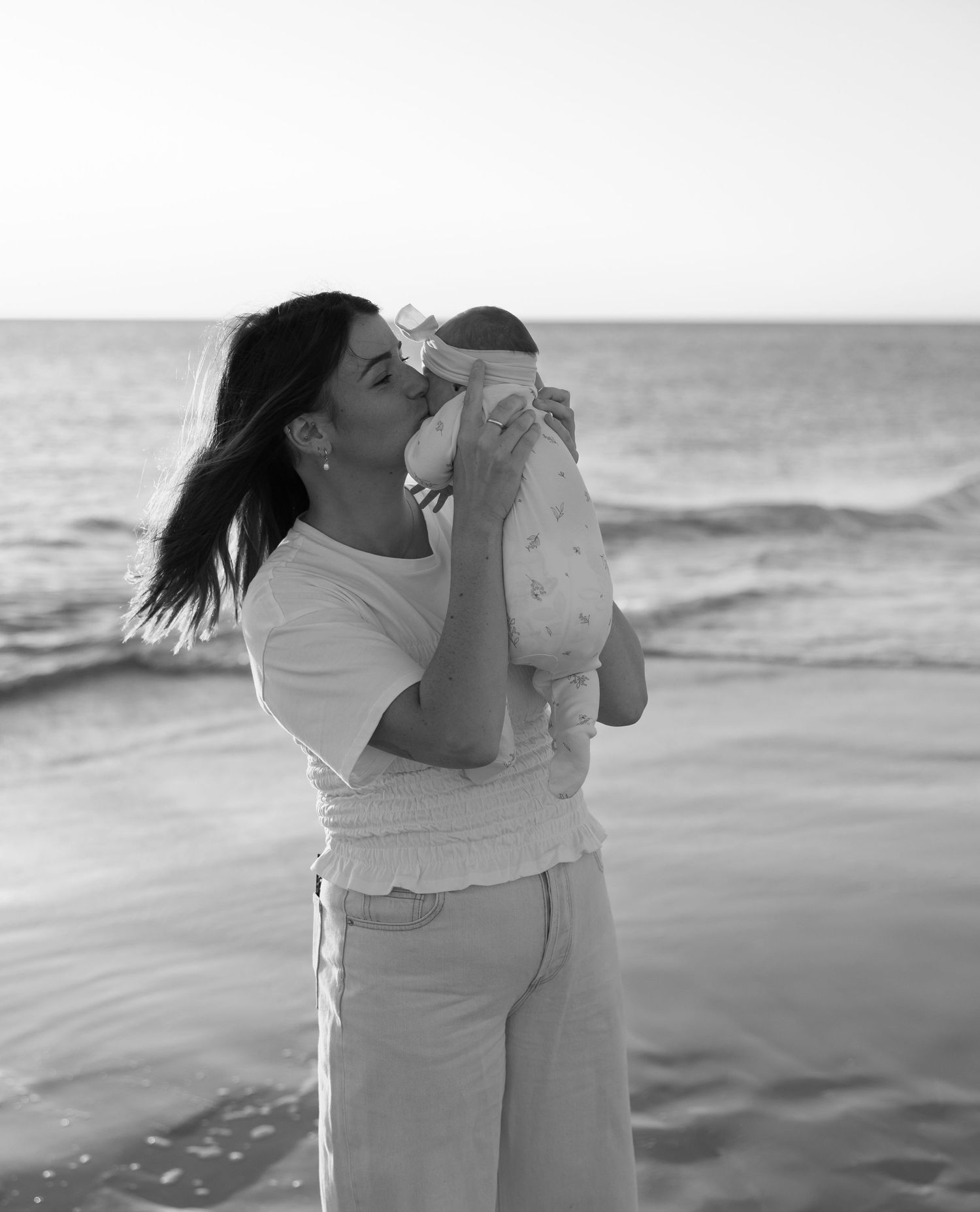 A tender moment captured in black and white as a mother holds her baby at the beach with waves in the background.
