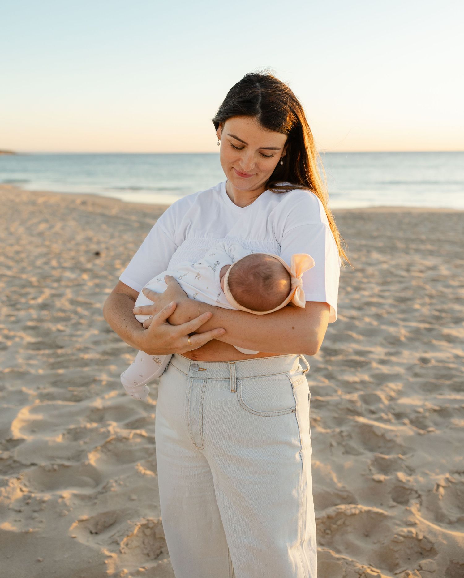 A tender moment captured at sunset on a sandy beach as a mother holds and gazes at her baby in soft natural lighting.