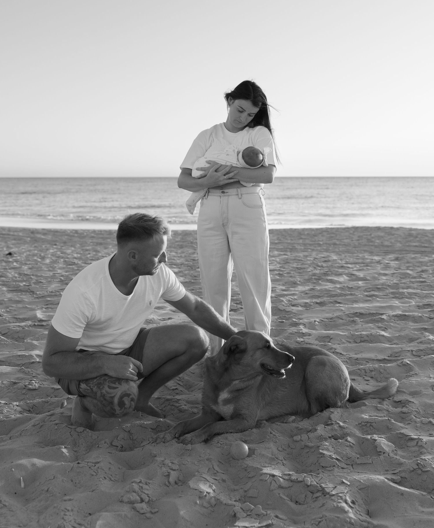 Black and white photo of a couple with their dog relaxing together on a sunny beach near the ocean.