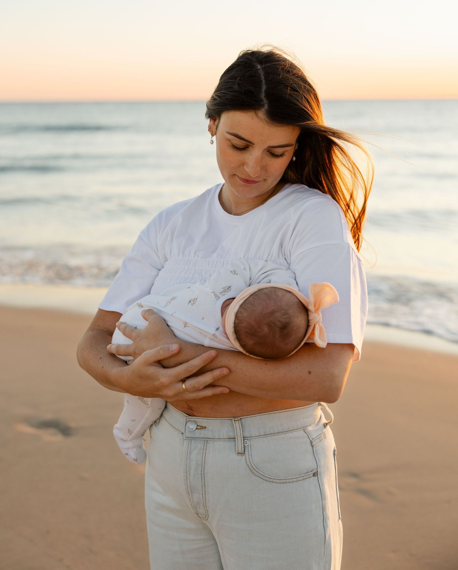A tender moment at sunset as a parent in white t-shirt and light jeans cradles a newborn on a peaceful beach.
