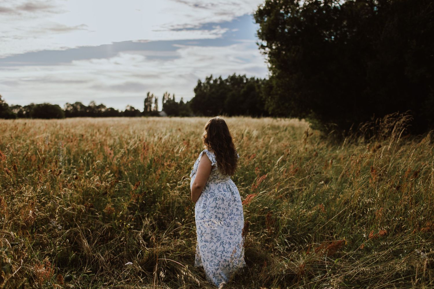 Woman in white floral dress standing alone in golden field at dusk.