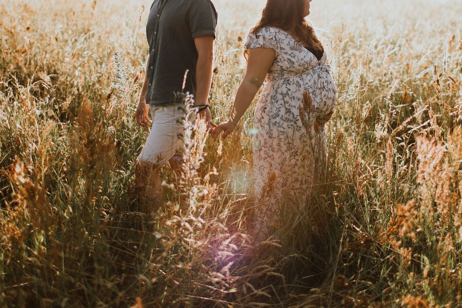 Couple holding hands while standing in tall golden grass during sunset.