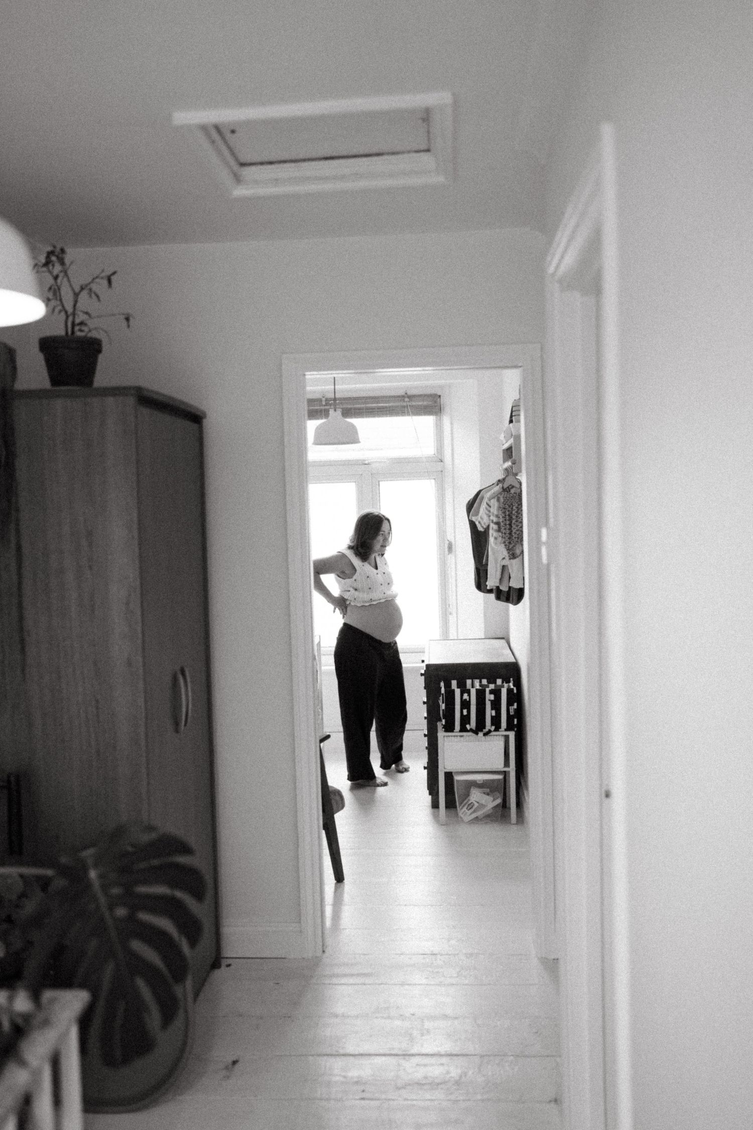 Black and white view down a hallway showing a figure in a doorway and wooden wardrobe against white walls.