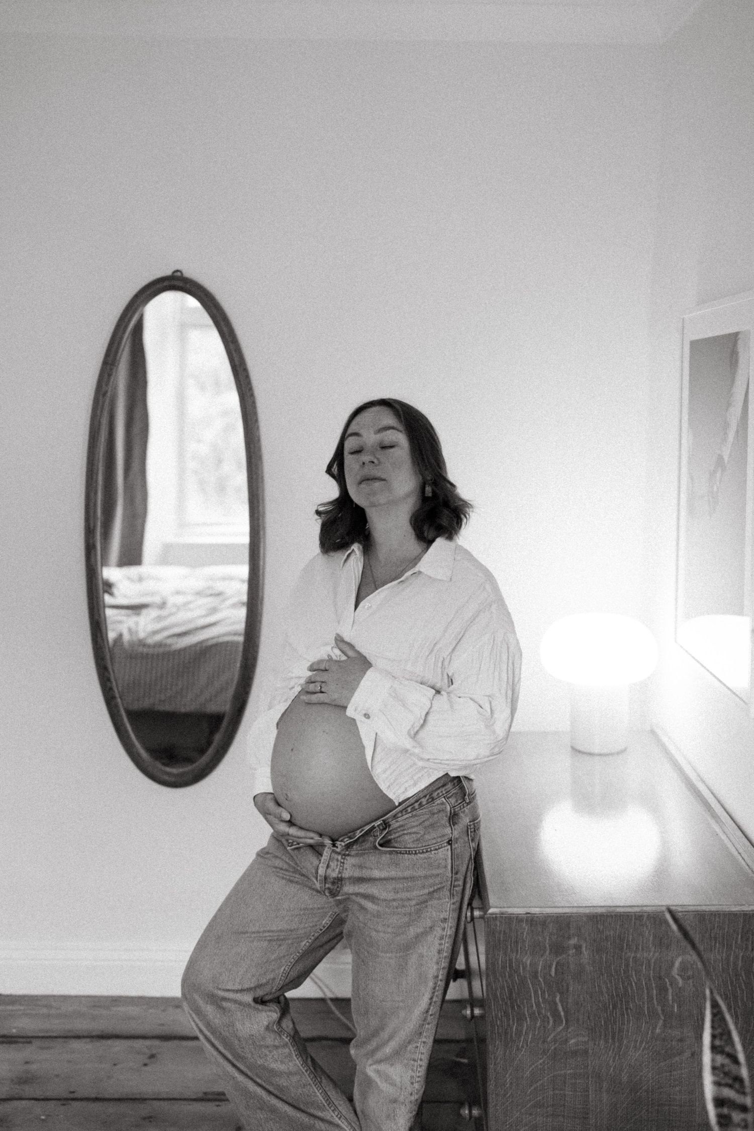 A black and white photo of an expectant mother in casual denim posing beside an oval mirror.