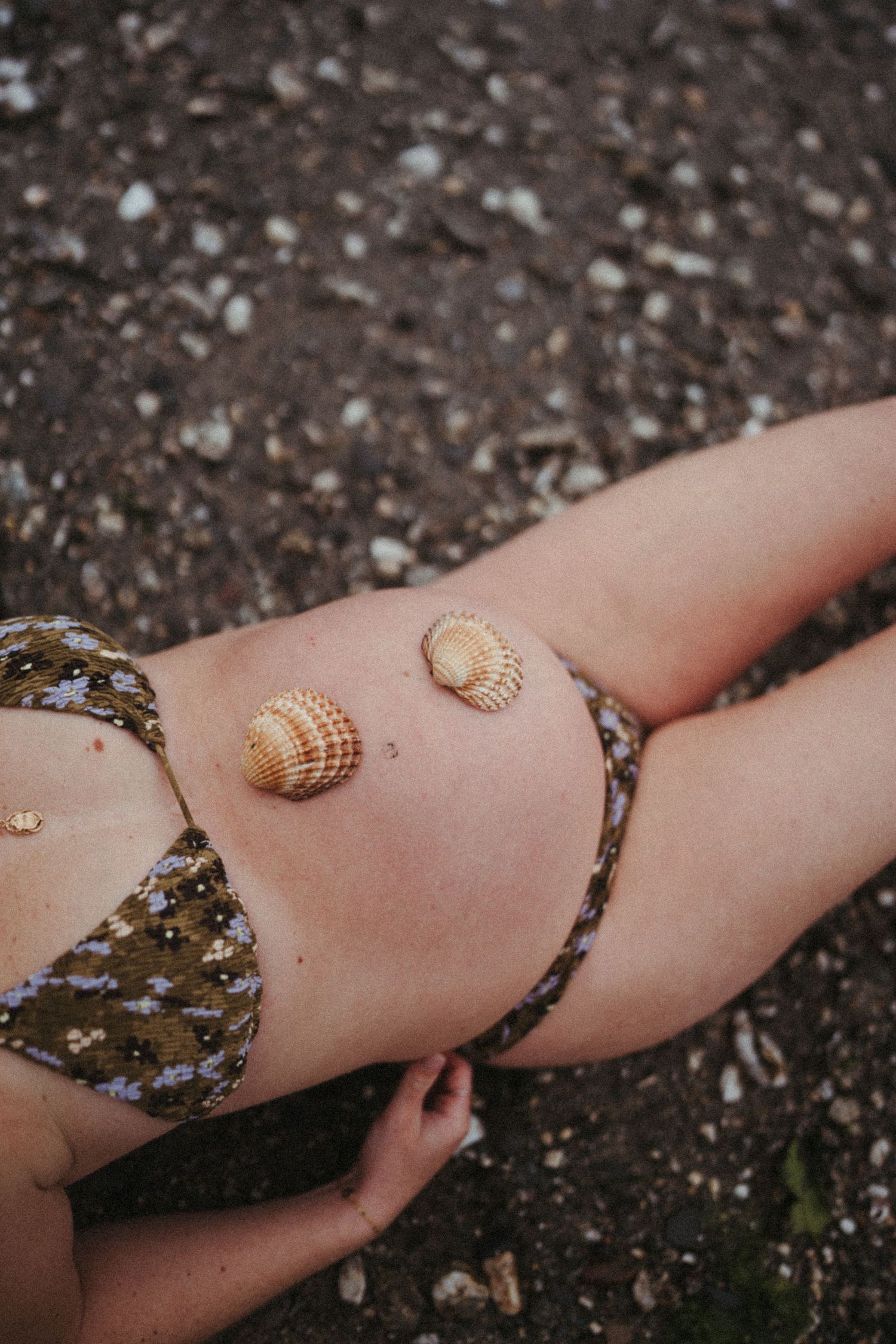 Close-up of a person wearing a shell bikini top on a dark sandy beach showing textured seashells against skin.