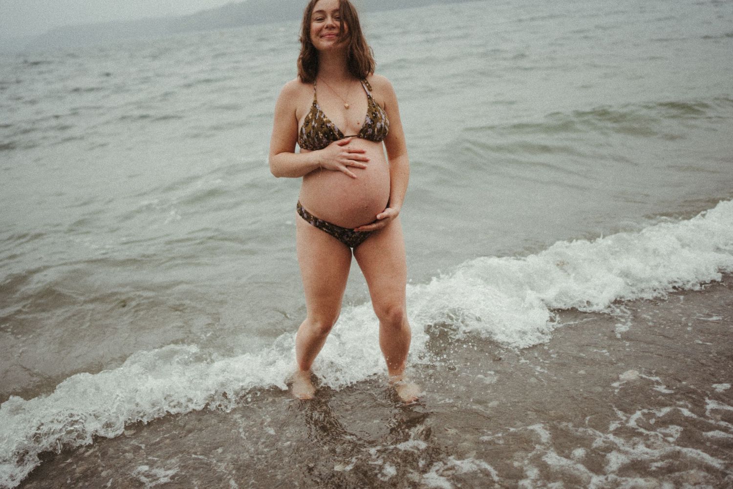 Expectant mother in a bikini stands at the ocean's edge as waves lap at her feet during a maternity photoshoot.