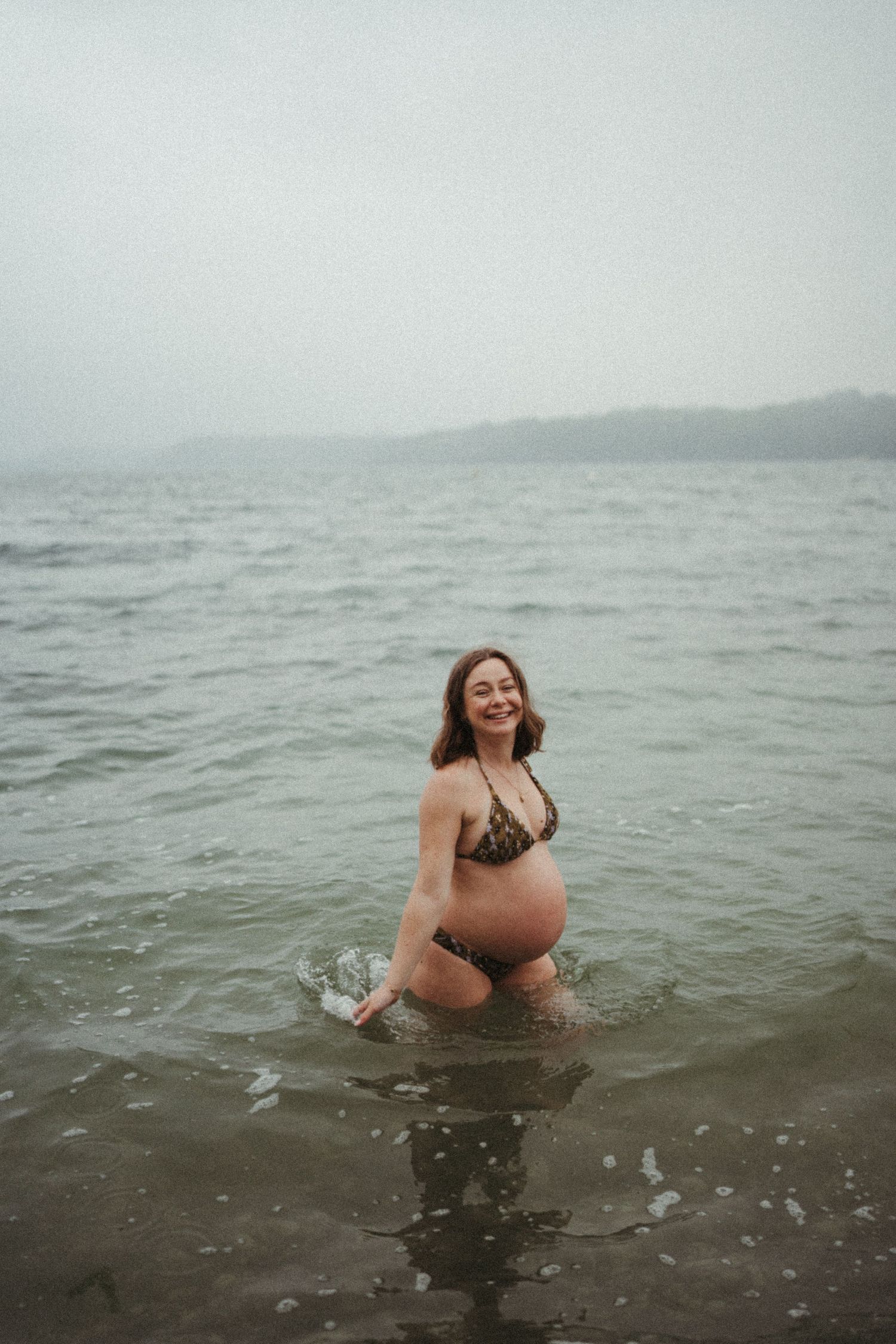 A pregnant woman in a bikini stands in gray ocean waters on a misty day enjoying a refreshing swim.