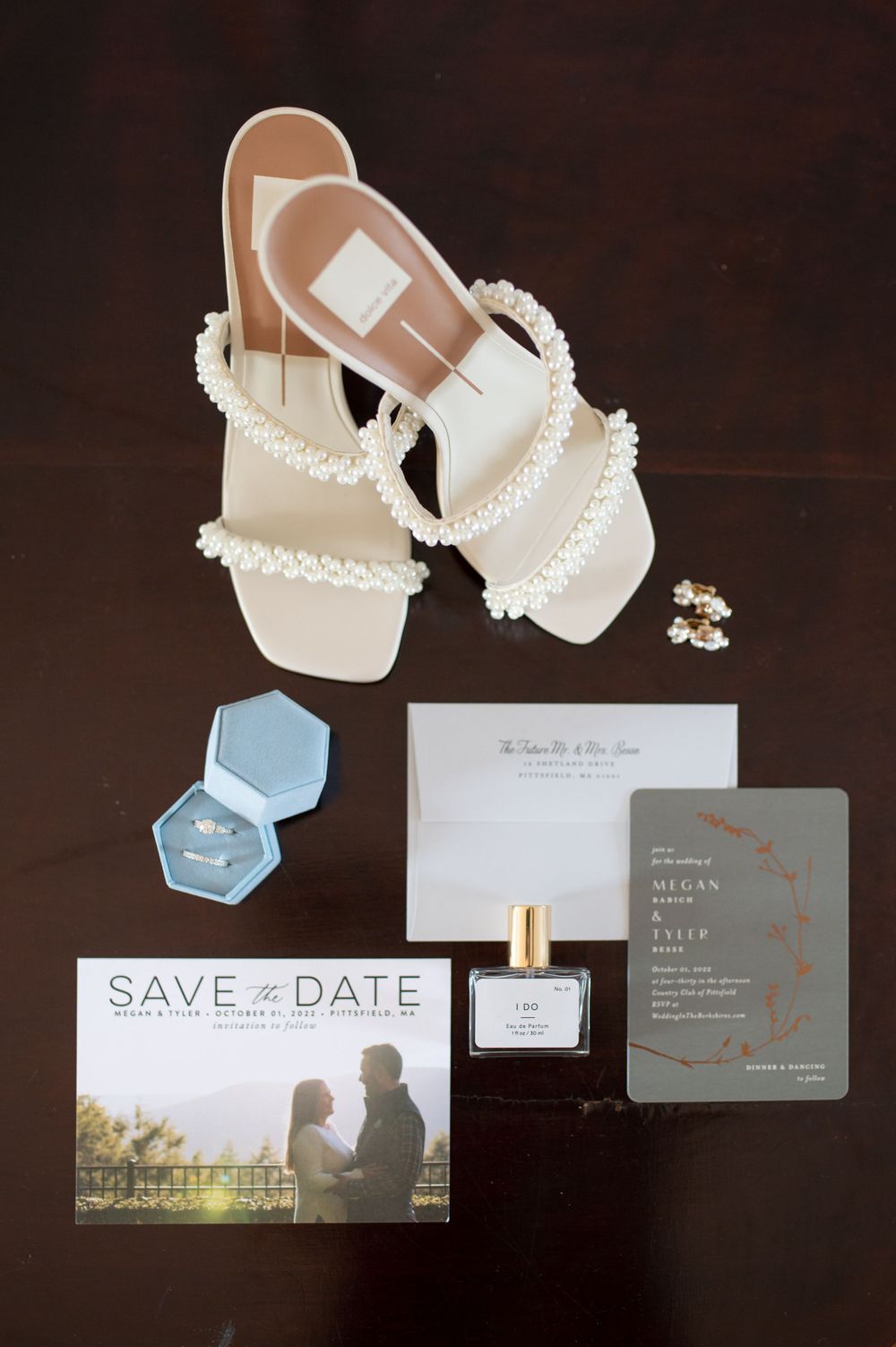 Wedding details flat lay with pearl sandals, save the date, perfume and ring boxes on dark background.