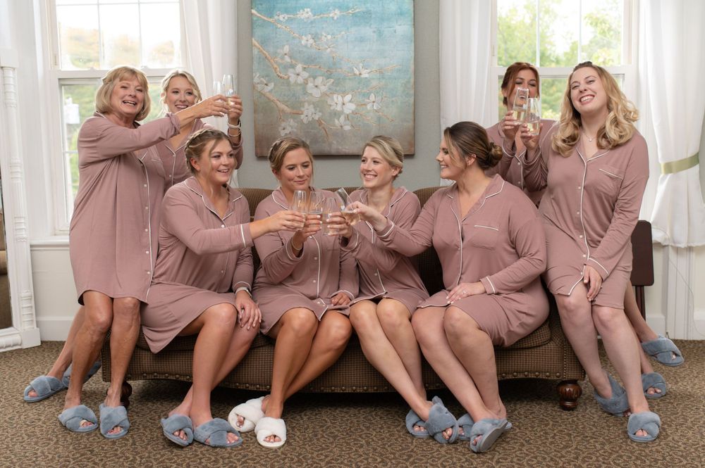 Bridesmaids in matching mauve robes toast with champagne while sitting together.