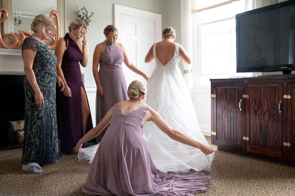 Wedding party helps adjust a flowing white bridal gown in a sunlit room.