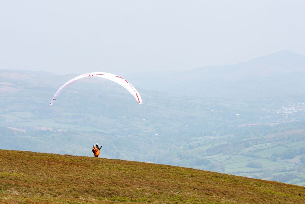 Paraglider with white wing launching from hilltop overlooking misty valley landscape.