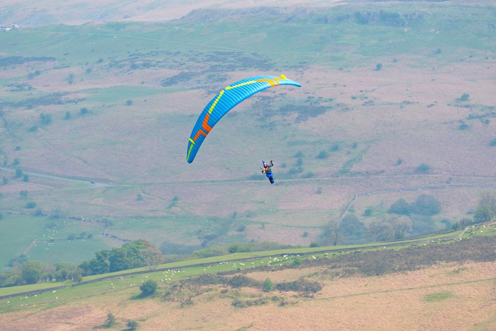Blue and yellow paraglider gliding over scenic mountain valley landscape.