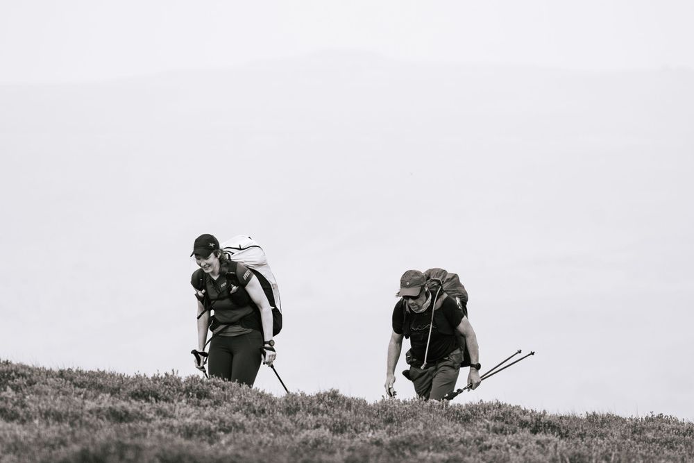 Two hikers with backpacks walking along ridge in black and white landscape.