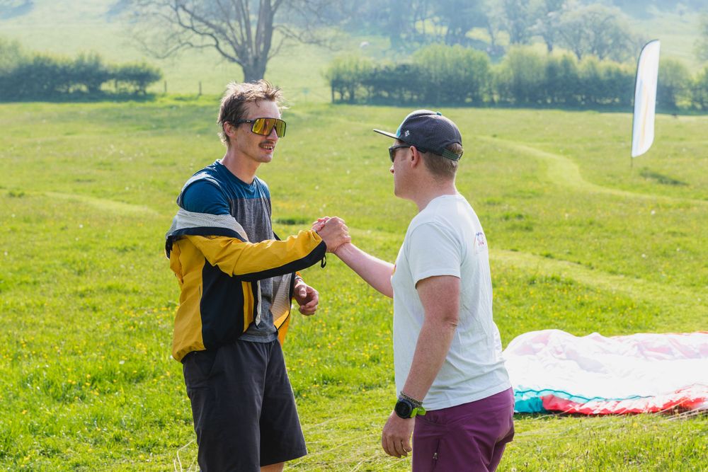 Two paragliders discussing on green field with equipment in background.