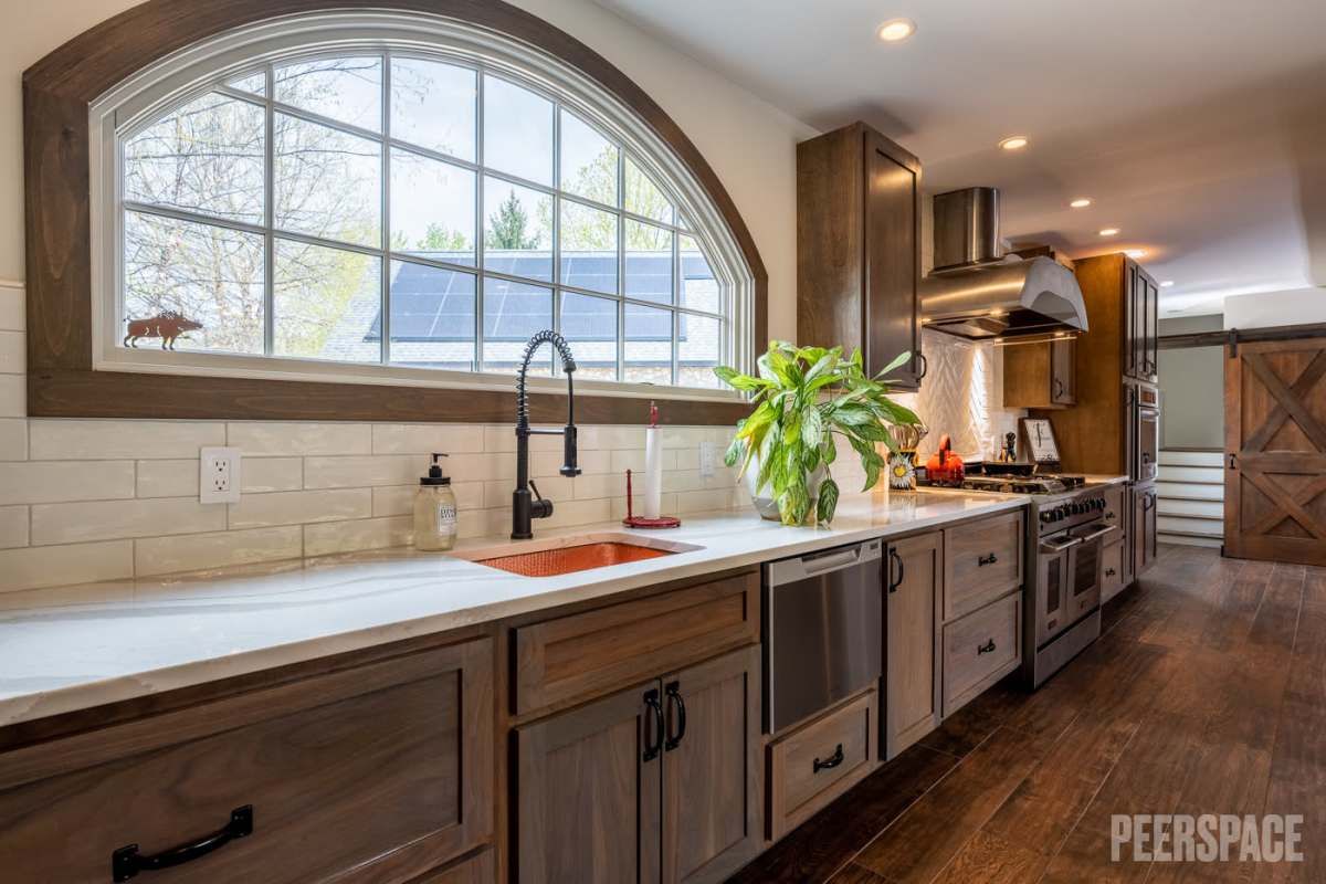 Modern rustic kitchen featuring dark wood cabinets, white countertops, and an arched window above the sink.