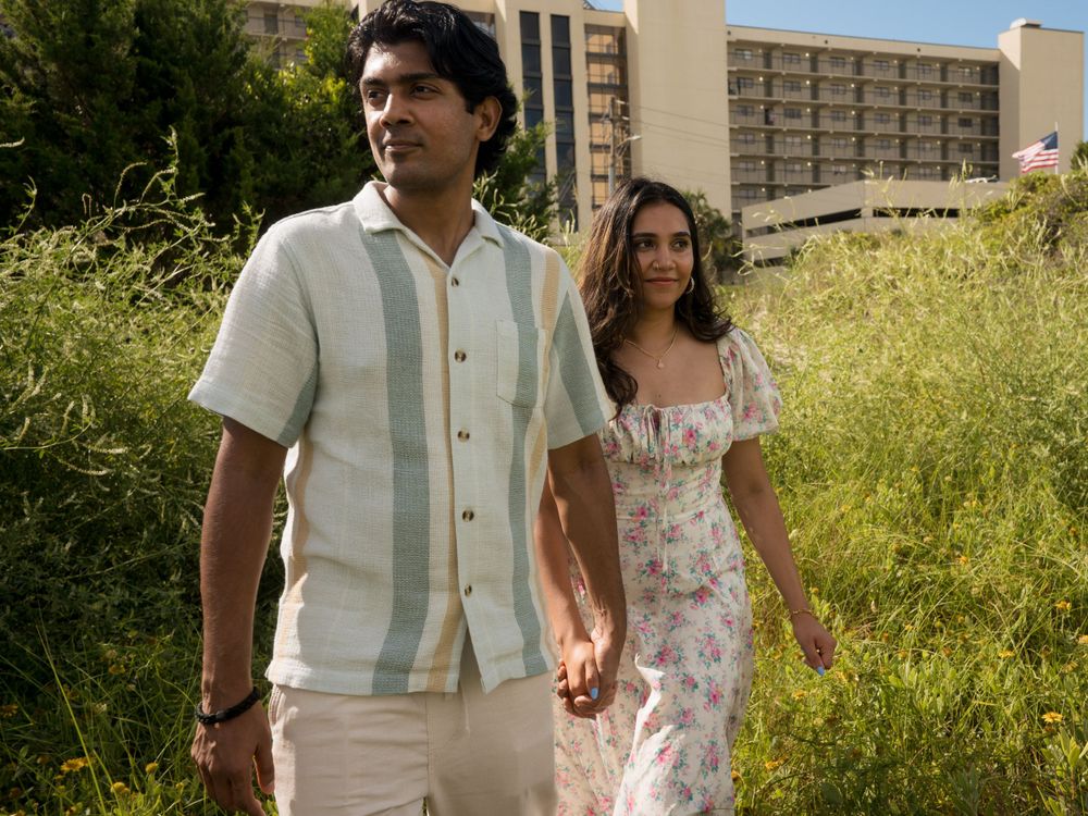 Two people walk together through a field of tall grass near modern apartment buildings.