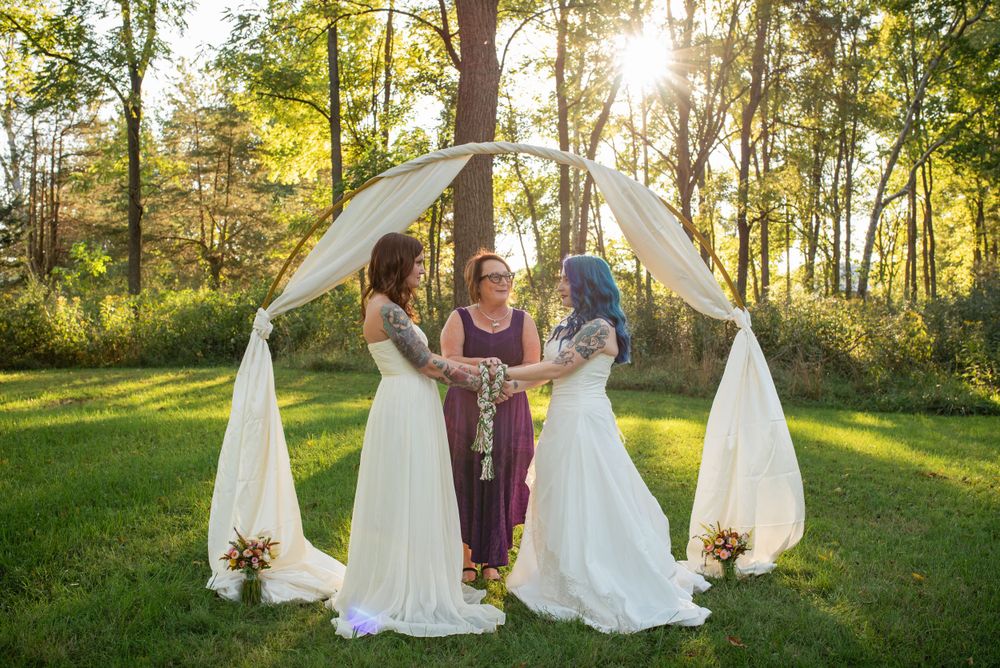 An outdoor wedding ceremony under a white fabric arch with sunlight streaming through trees.