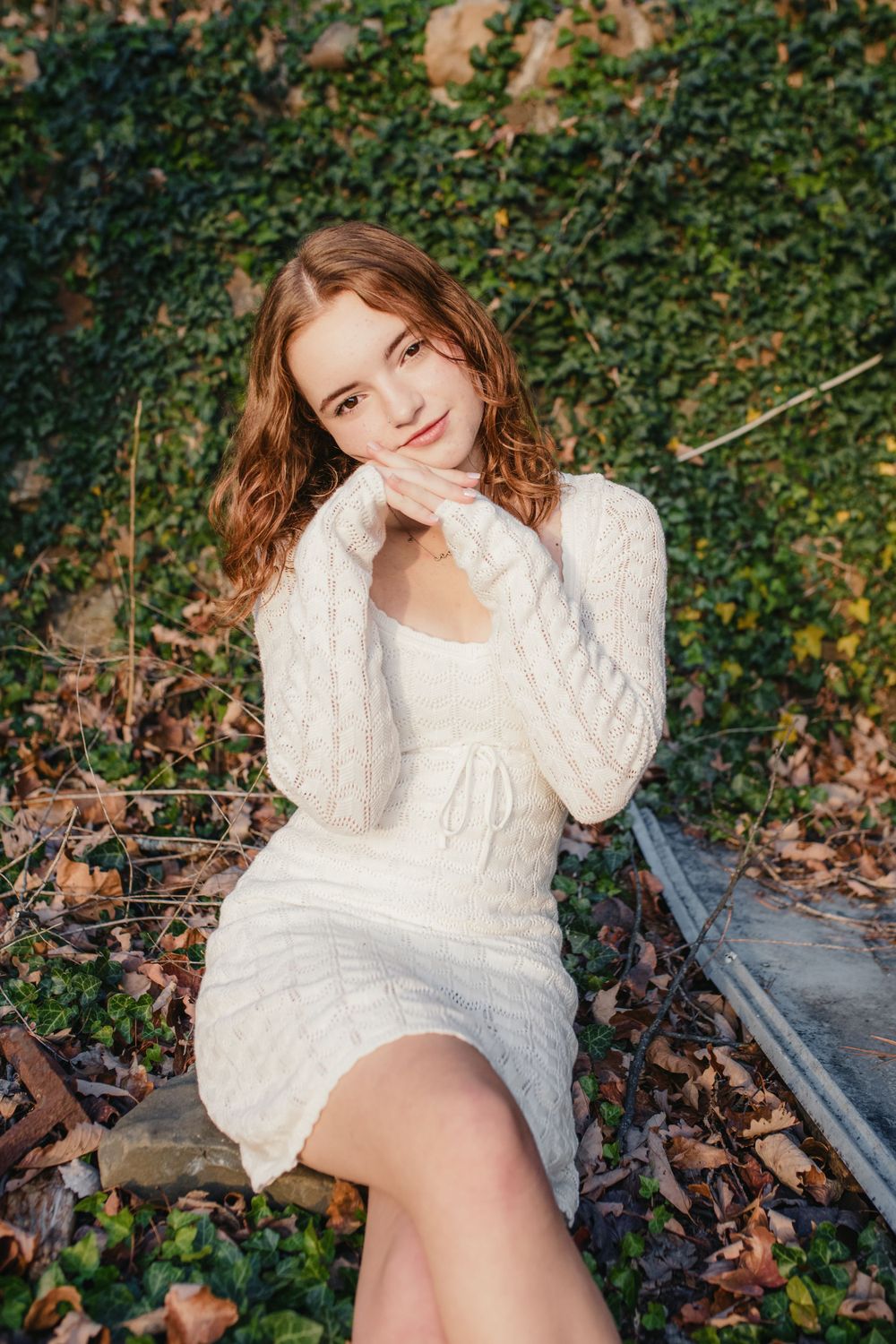A portrait in a white knit dress against an ivy-covered wall background.