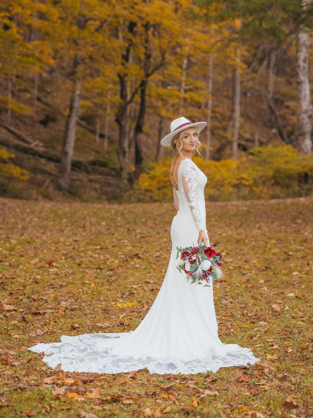 A bride in a lace gown poses in a fall forest setting with autumn foliage.