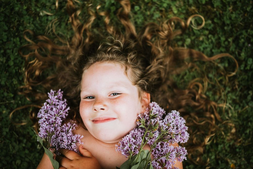 A whimsical portrait with purple lilac blooms and curly hair against green foliage.