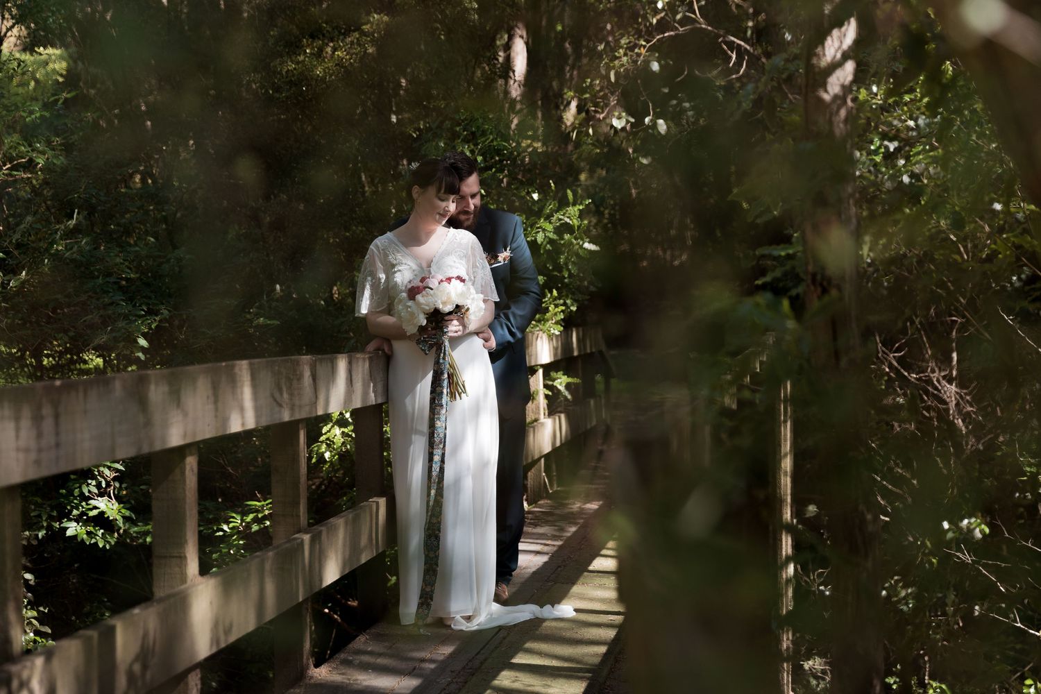 Wedding couple stands together on wooden boardwalk surrounded by lush greenery.