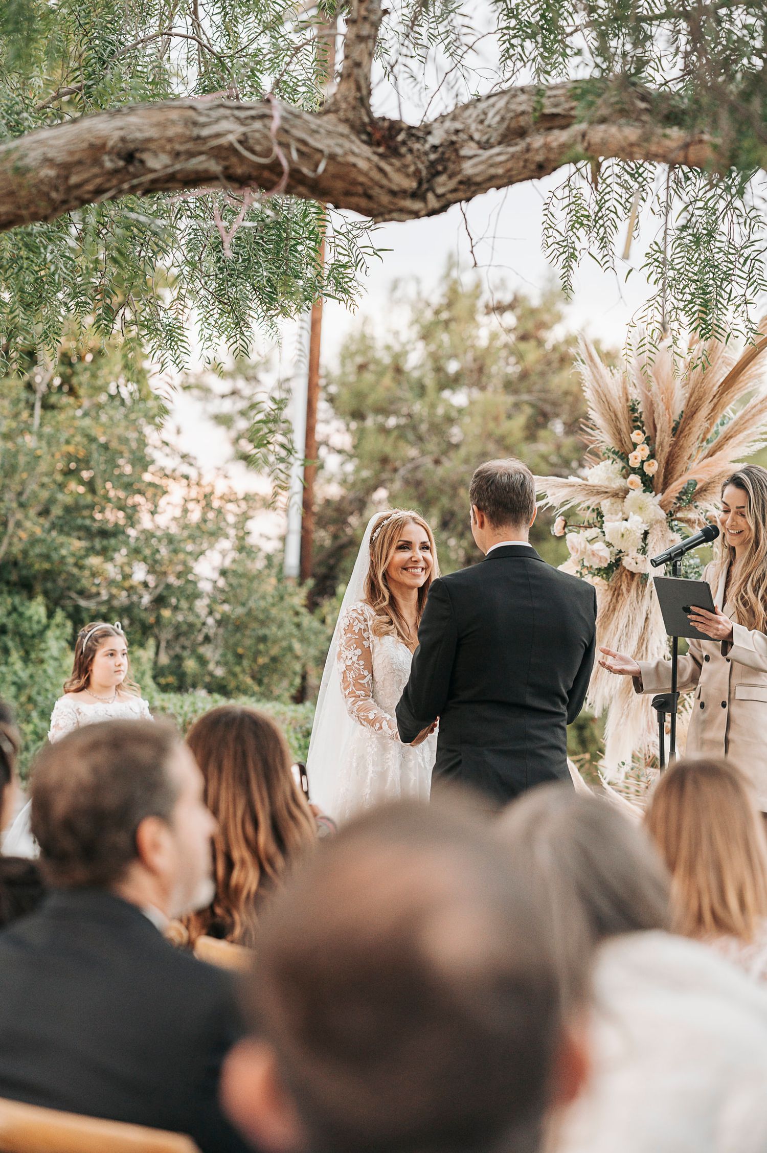 An outdoor wedding ceremony under a twisted tree branch with guests watching the exchange of vows.