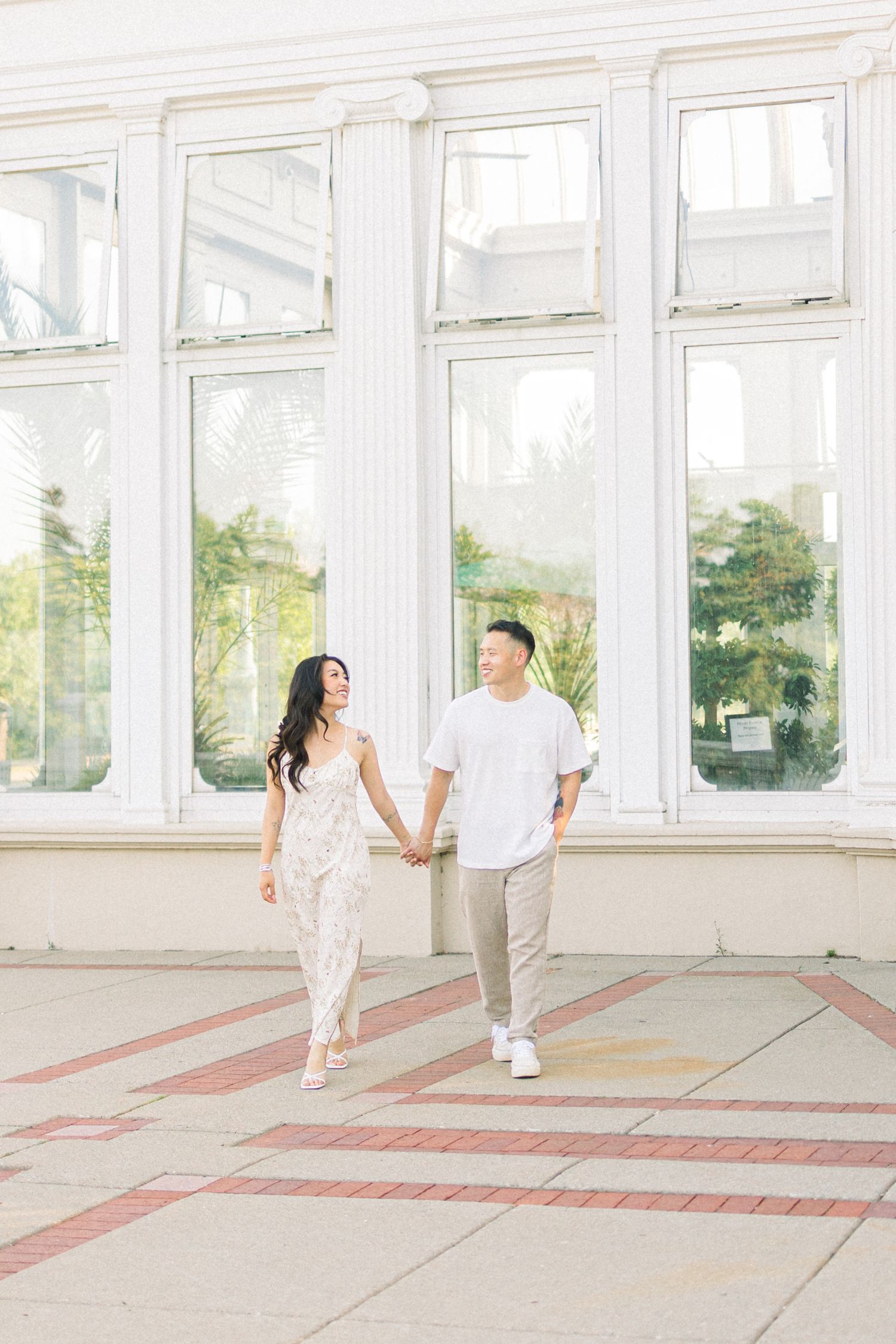 Couple walking hand in hand along bright sunlit corridor with large windows.