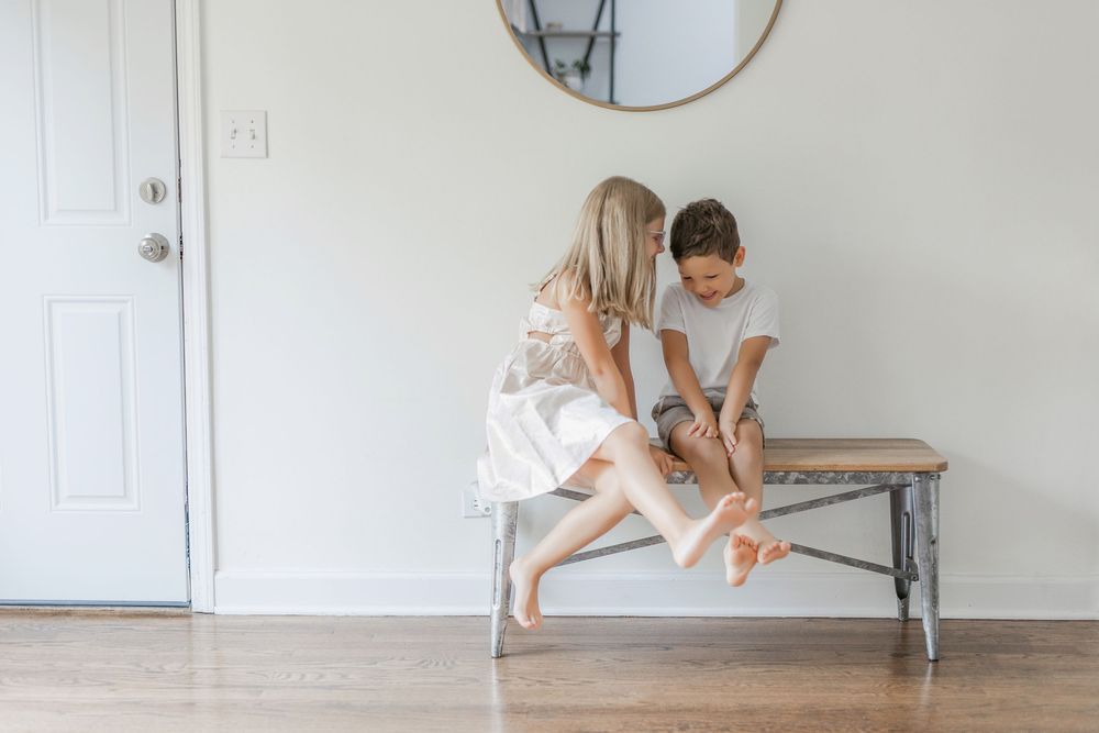 Two people in white outfits sit together on a wooden bench against a light gray wall with a circular mirror.
